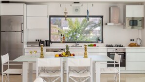 A modern kitchen with white cabinetry, stainless steel appliances, a large window with a view of greenery, and a dining area with a glass table and chairs. - Villa Setara is a rare beach front property located in Kalkan (Kalkan)