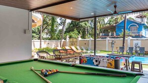 Une table de billard et une table de baby-foot sont installées dans un espace extérieur abrité, agrémenté de fauteuils et offrant une vue sur un bâtiment bleu et des palmiers.