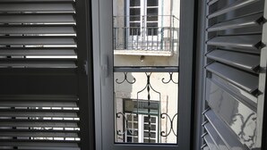 Vista a través de una ventana con persianas metálicas, mostrando un balcón con barandilla y la fachada de un edificio con ventanas.