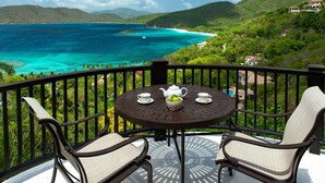 Un balcon avec une table, des chaises et une vue sur une mer turquoise et des collines verdoyantes.