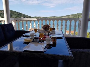 Een eettafel met ontbijtartikelen en een glas sinaasappelsap op een balkon met uitzicht op een waterlichaam en heuvels.