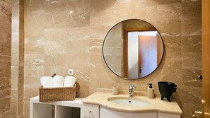 Ein Badezimmer mit Marmorwand, einem runden Spiegel, einem Waschbecken, einem Korb mit Handtüchern und einem schwarzen Seifenspender.