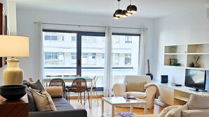 Un salon moderne comprenant un canapé, un fauteuil, une table basse et un téléviseur. On y trouve également un coin repas avec des chaises et une table. La pièce est dotée de grandes fenêtres et d’un luminaire au plafond.