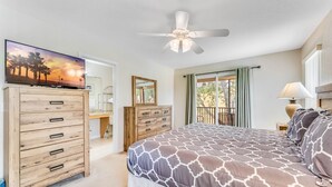 Ein Schlafzimmer mit einem Bett, einem Schminktisch, einem Fernseher, einem Deckenventilator und einem Balkon mit Vorhängen.