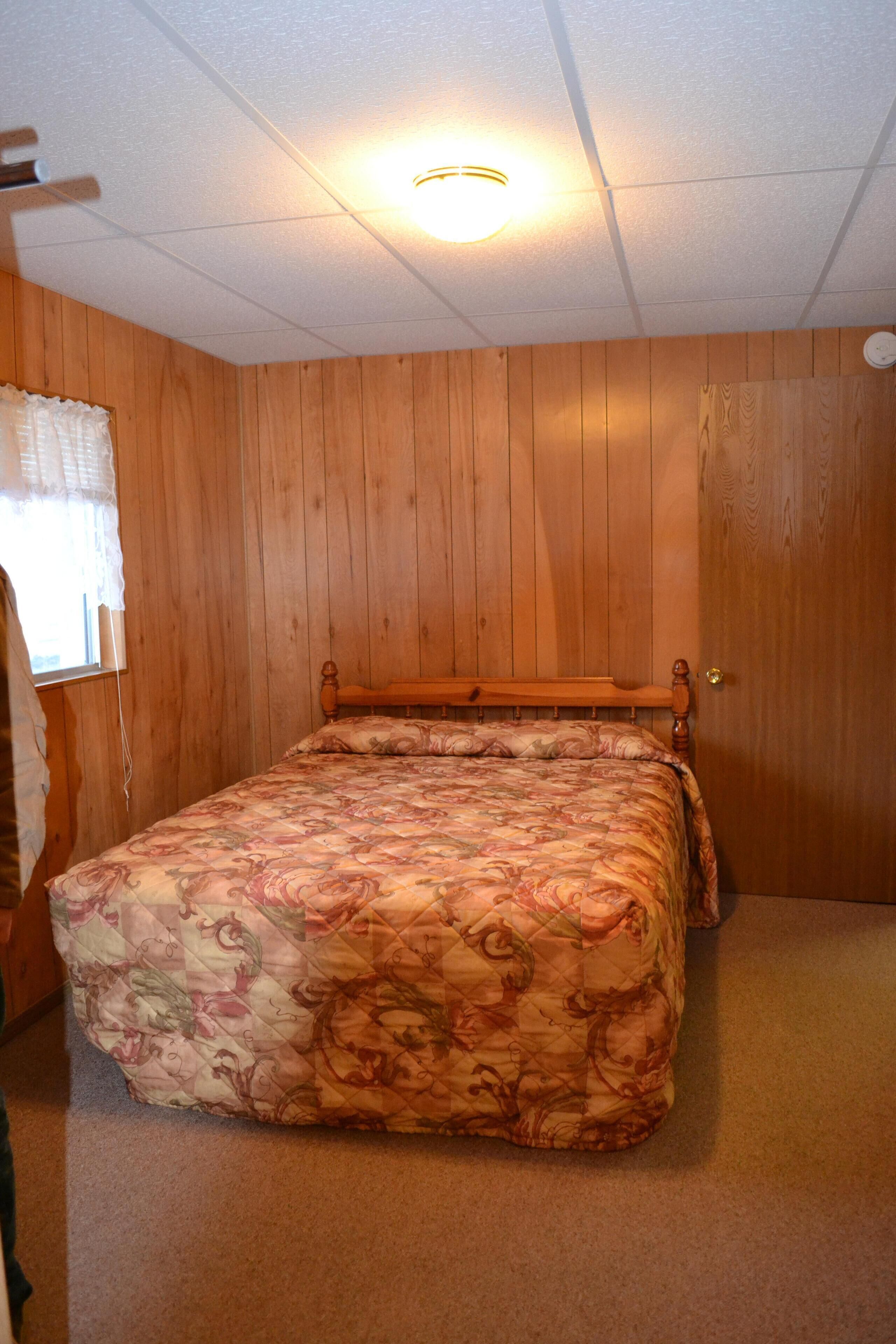 Une chambre avec des murs lambrés, un lit recouvert d’une couvre-lit à motifs floraux, un ventilateur de plafond et une fenêtre avec des stores.