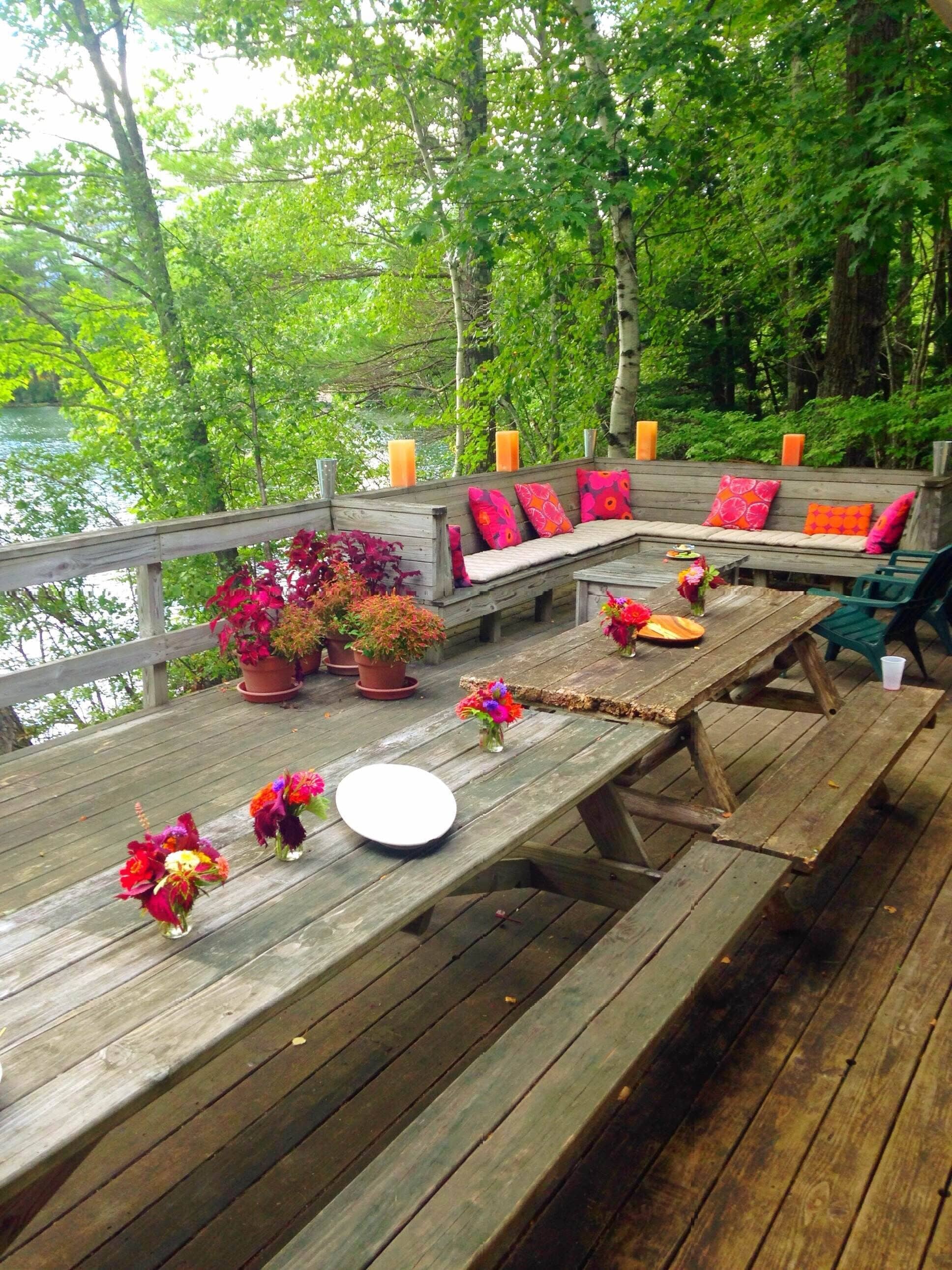 A wooden deck with a bench and table, surrounded by lush greenery and a body of water.
