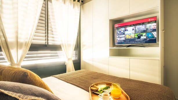 Hotellihuone, jossa on sänky, tarjotinalus aamiaisella, televisiossa uutislähetyksiä ja ikkunassa verhot.