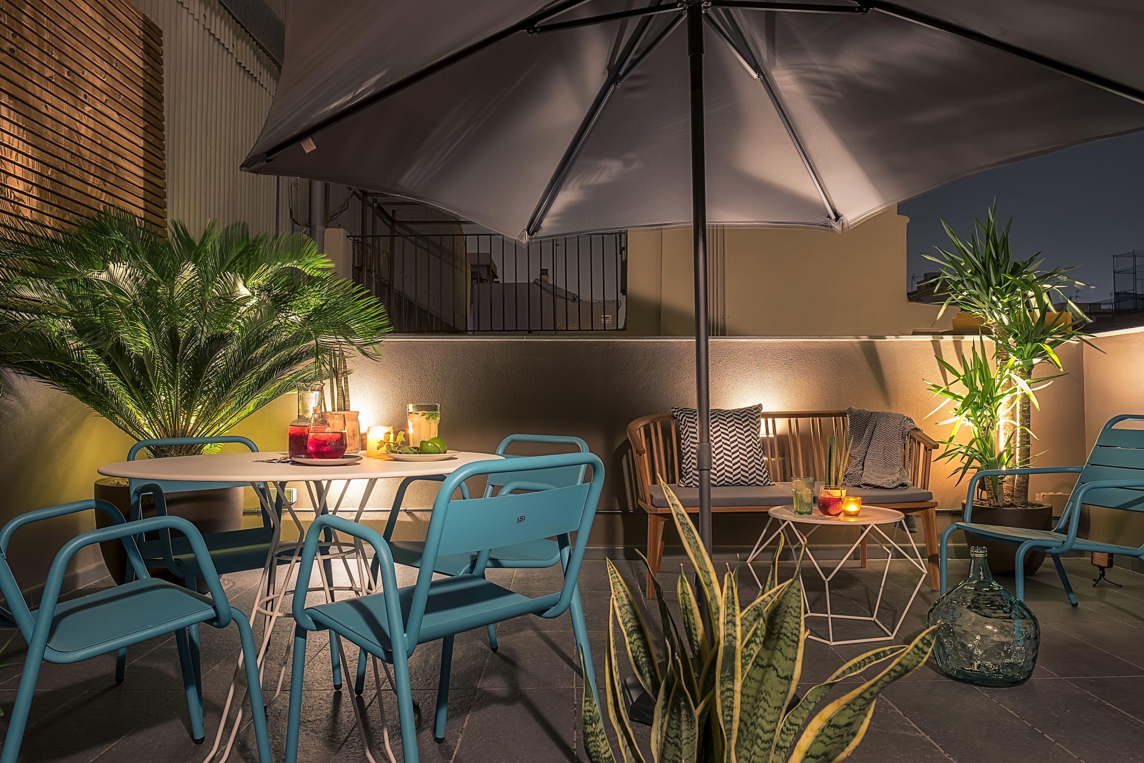 Een terras buiten met een tafel, stoelen, een grote parasol en potplanten.