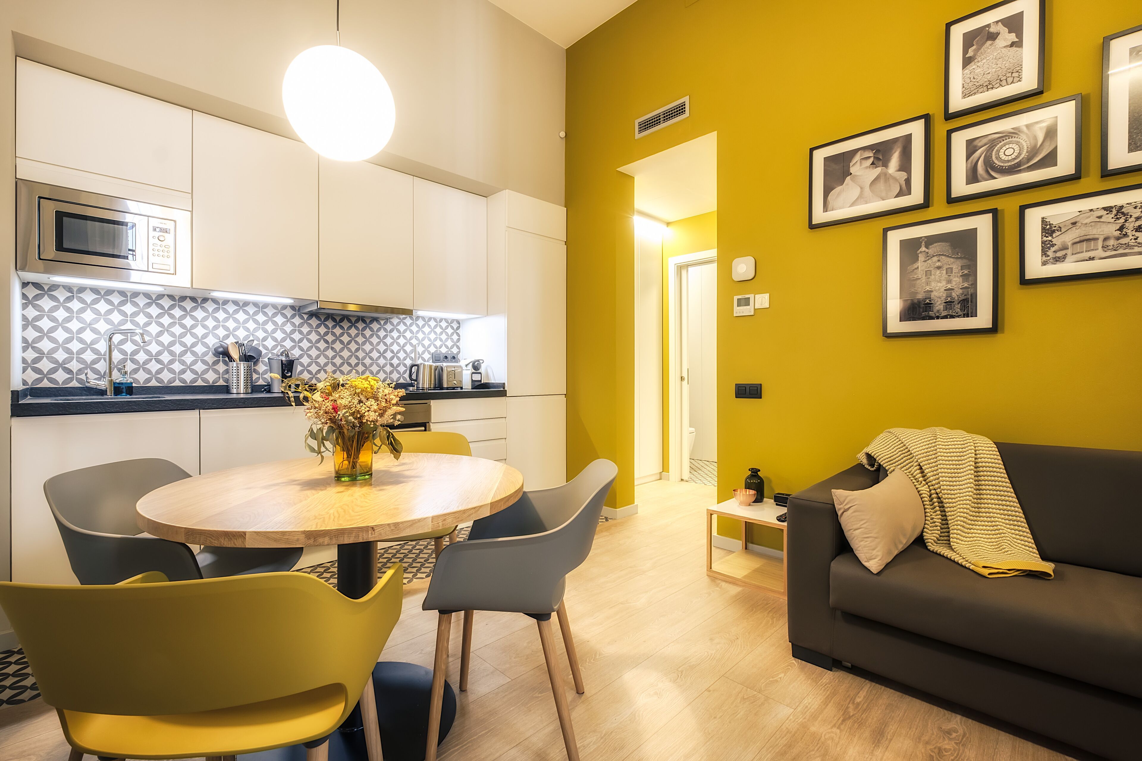 Een moderne keuken met een magnetron, een eettafel met stoelen en een gele wand met ingelijste foto's.