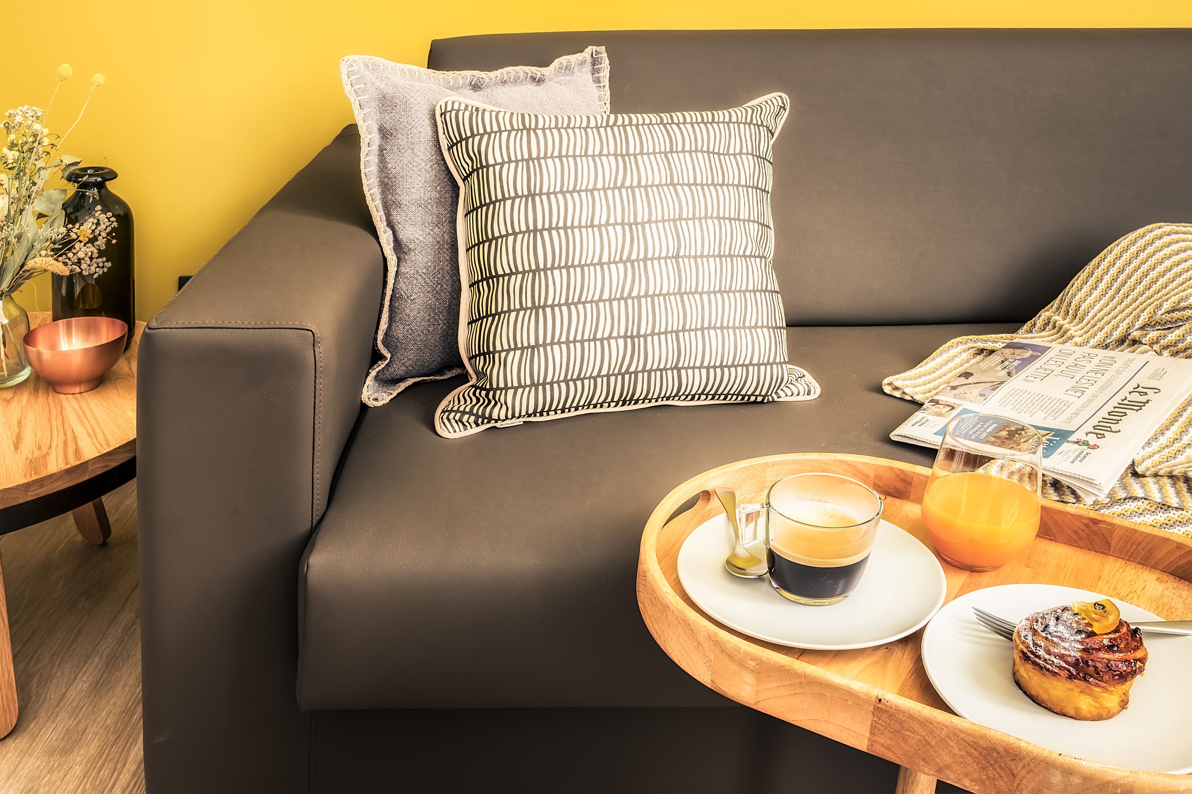 Een moderne woonkamer met een donkere bank, een houten bijzettafeltje, een dienblad met een kop koffie, een glas sinaasappelsap en een bord met gebak.
