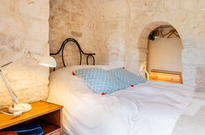 A bedroom with a stone wall, a bed with white bedding and blue patterned pillows, a bedside lamp, and a wooden nightstand. - Trulli e Masseria Ianella (Locorotondo (Bari))