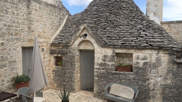 Um edifício de pedra com telhado em tijolos, uma porta e janelas. Possui áreas de estar ao ar livre com cadeiras e uma mesa, além de plantas em vasos.