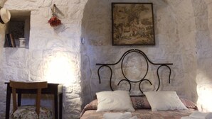Quarto com parede de pedra, cama com roupa de cama branca, uma cadeira de madeira e um quadro.