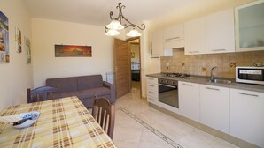 A compact kitchen with white cabinets, a microwave, and a gas stove. There is a dining table with a checkered tablecloth and a bowl of fish. A brown sofa is against the wall, and a painting hangs above it. - Apartment with Garden View (Trecastagni)