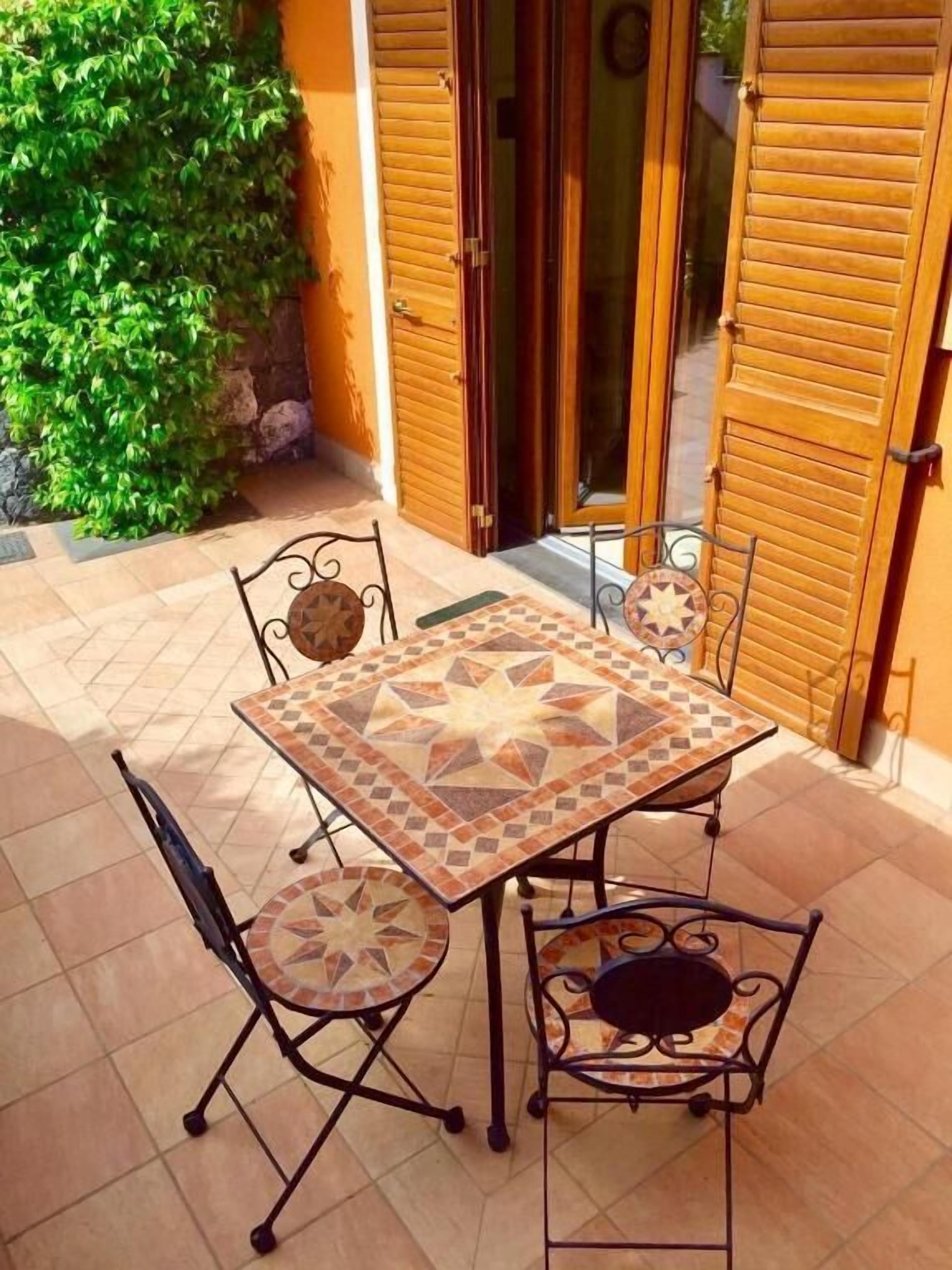 Un patio con un tavolo piastrellato e quattro sedie, circondato da persiane in legno e un arbusto verde.