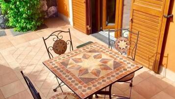 Un patio con un tavolo piastrellato e quattro sedie, circondato da persiane in legno e un arbusto verde.