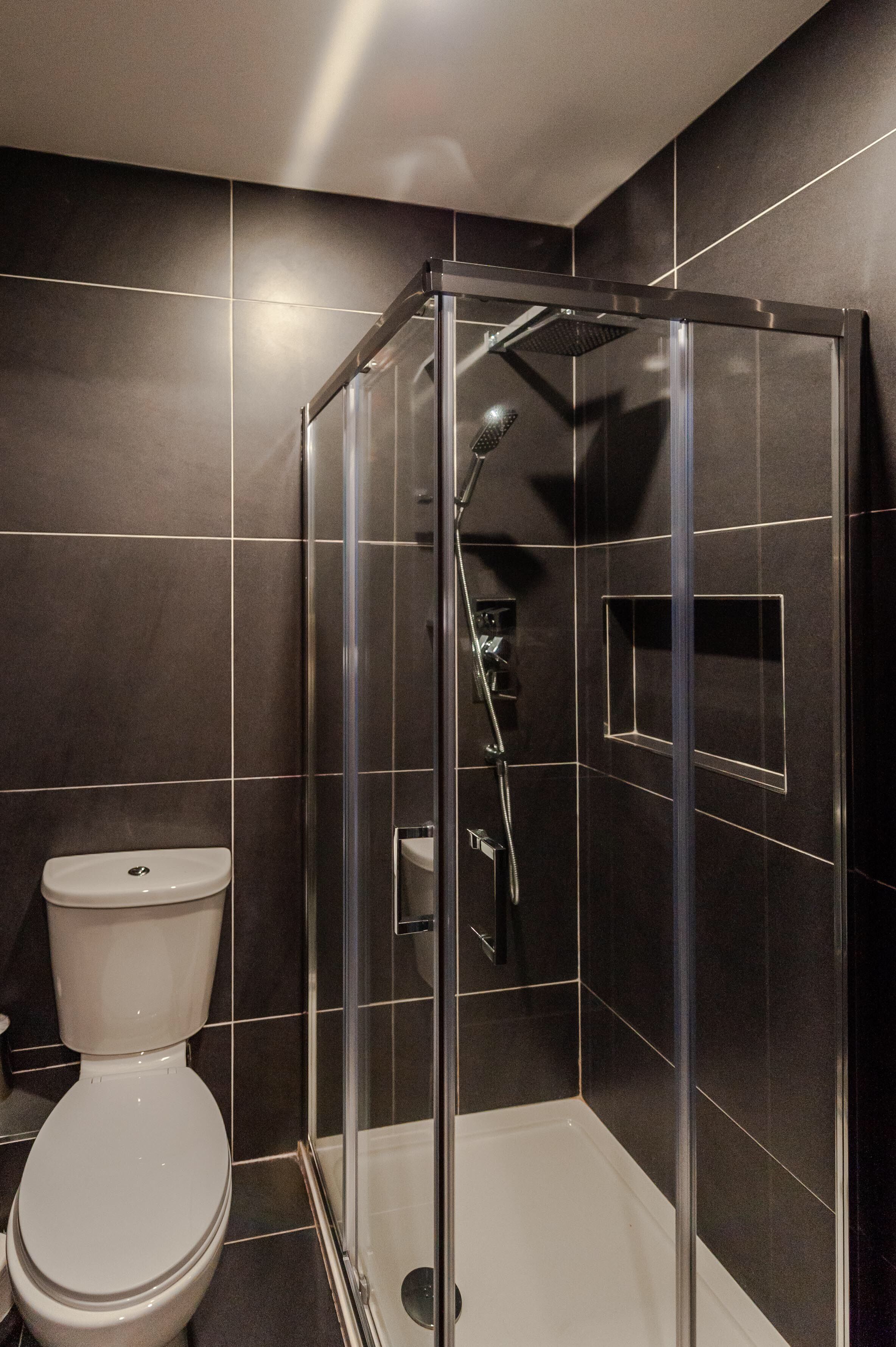 A modern bathroom with a glass-enclosed shower, a white toilet, and dark tiled walls.