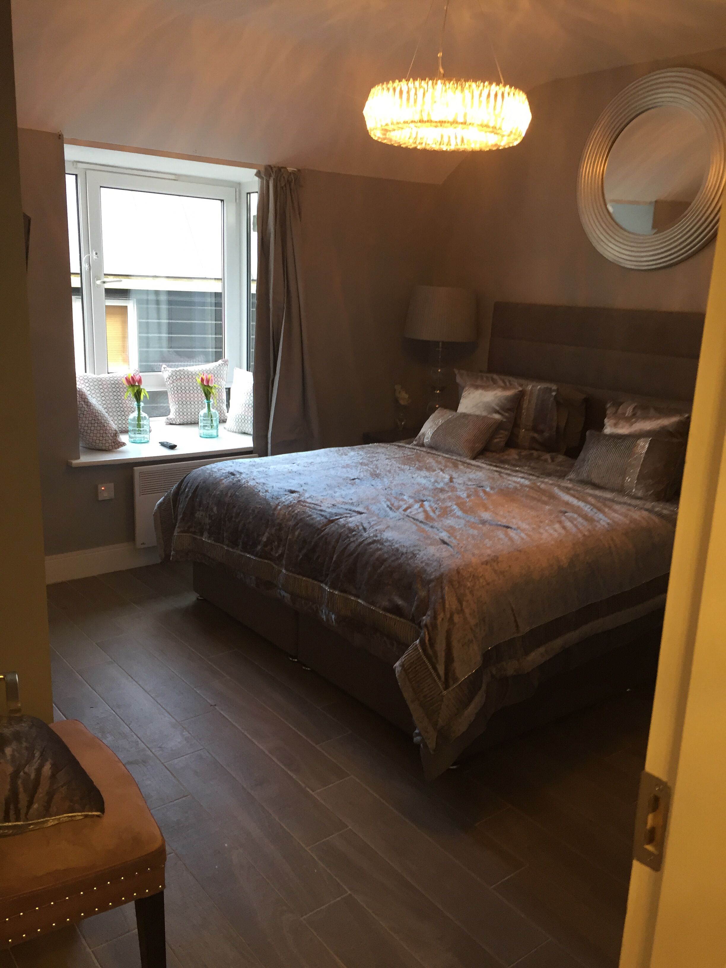 A bedroom with a large bed, a bedside table, a lamp, a window with curtains, and a chandelier.