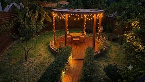 A wooden deck with a round table and chairs, surrounded by a garden with various plants and a wooden fence. - Minoa Downtown  Garden villa (Κουνουπιδιανά)