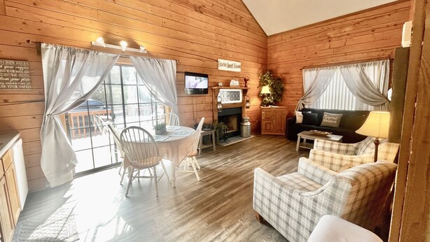 A cozy cabin interior with wooden walls, a dining area with a table and chairs, a fireplace, a television, and a sofa.