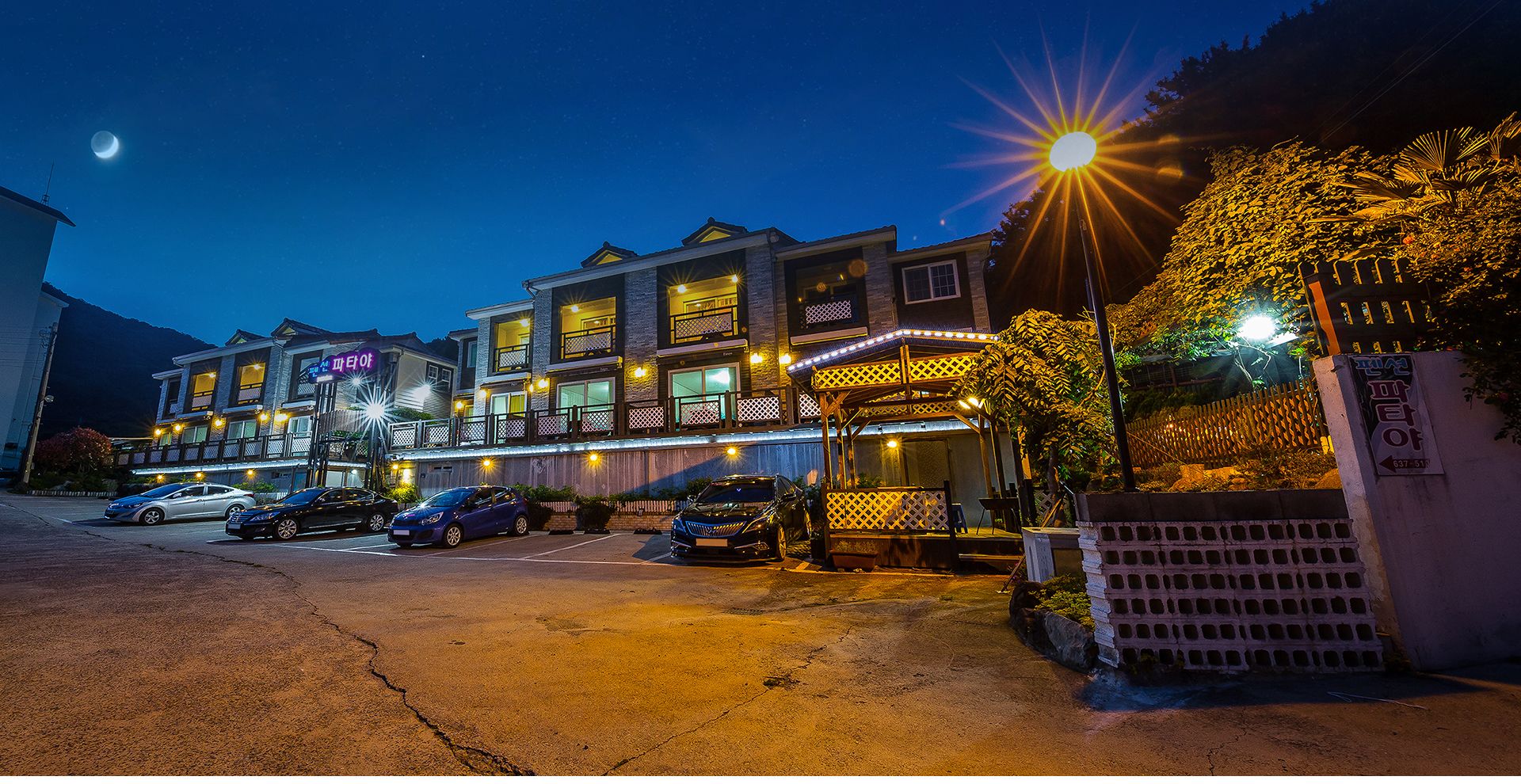 Um hotel com vários andares, uma fileira de varandas, uma área de estar coberta ao ar livre e um estacionamento com vários carros.