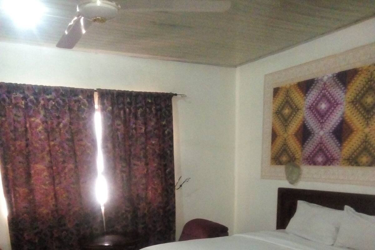 Quarto de hotel com uma cama, uma cadeira, uma mesa pequena e um ventilador de teto.