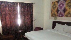 A hotel room with a bed, a chair, a small table, a ceiling fan, and a decorative wall hanging. - Pridemark Hotel (Abuja)