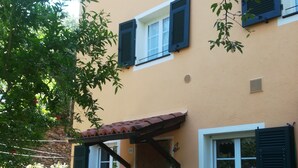 Un bâtiment à deux étages avec une façade jaune, des volets verts et un toit recouvert de tuiles. On y trouve des plantes en pot et un banc en bois à l’extérieur.