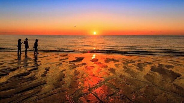 Escena de playa con atardecer, tres personas de pie sobre la arena y el océano al fondo.