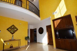 Uma sala de estar ampla com teto alto, varanda com guarda-corpo, TV de tela plana e um objeto decorativo fixado na parede.