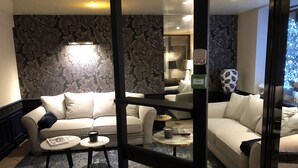 Un salon moderne avec un canapé, une table basse et une petite table d’appoint. Les murs sont recouverts d’un papier peint à motifs et une cloison en verre sépare l’espace.