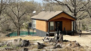 Une cabane en rondins avec un petit porche et une table de pique-nique, entourée d’arbres et bordée par un plan d’eau.