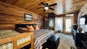 Une chambre située dans une cabane en rondins, comprenant un lit, un bureau, une chaise, un ventilateur de plafond, un escalier et une porte donnant sur l’extérieur.