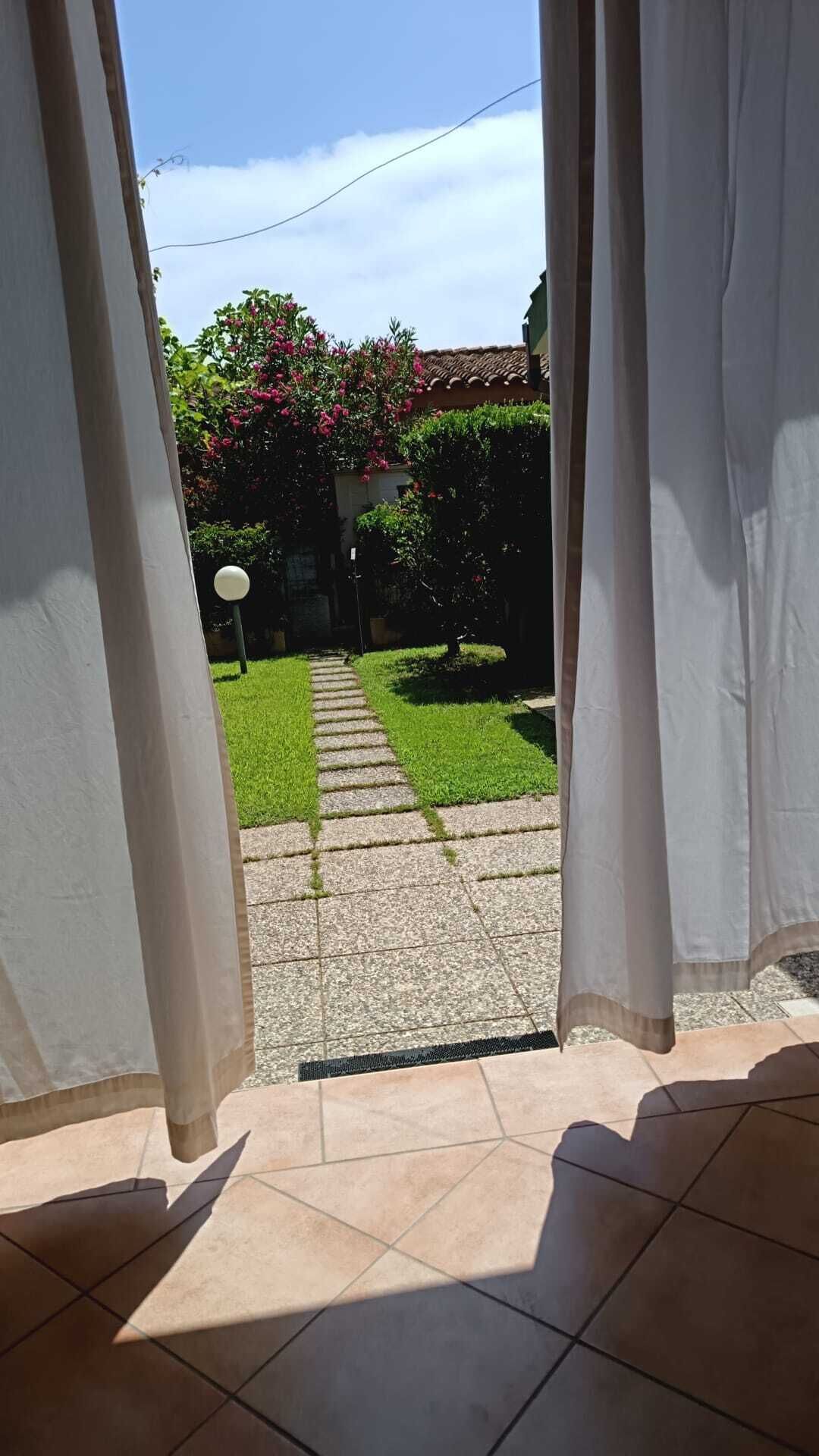 Uitzicht door een deuropening op een binnenplaats met een stenen pad, groen gras en een boom met roze bloemen.