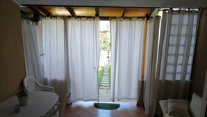 Une pièce avec un sol carrelé, une table blanche avec des chaises, un canapé et une vue sur un jardin, visible à travers une porte munie de rideaux.