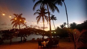 Un espace de restauration situé au bord de la plage, avec des tables et des chaises, des palmiers et des guirlandes lumineuses.
