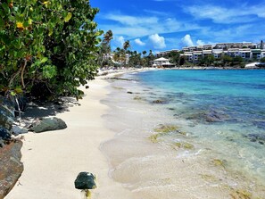 A beach with clear turquoise water, sandy shore, and a row of buildings in the background.