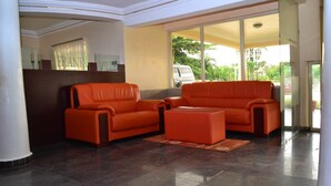 A lobby with two orange sofas and a matching ottoman, a large mirror, and a view of a van outside through large glass doors. - Frankville Hotel (Abuja)