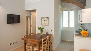 A dining area with a wooden table and chairs, a vase of flowers, and a kitchen area with a stone archway and a fruit bowl. - La Corte di Zizi-apartment 'Riva': when grandparents travel with you (Cernobbio)