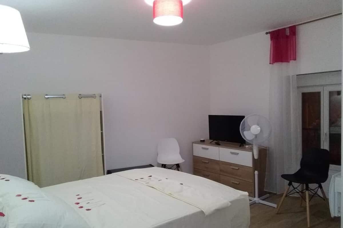 A bedroom with a bed, a desk, a chair, a TV, and a fan.