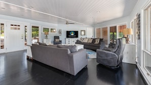 Un salon spacieux avec un grand canapé gris, un fauteuil en cuir noir et une table basse blanche. Un téléviseur à écran plat est fixé au mur, ainsi qu’un ventilateur de plafond et une bibliothèque contenant divers objets.