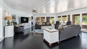 Un vaste salon doté d’un parquet en bois foncé, d’un plafond peint en blanc et de grandes fenêtres offrant une vue sur la verdure environnante.