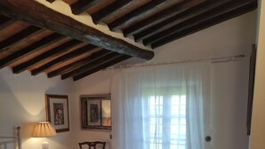 A bedroom with a bed, a chair, a nightstand, a lamp, a window with curtains, and a small table with a vase. - Casa Crevole in Val di Merse (Murlo)