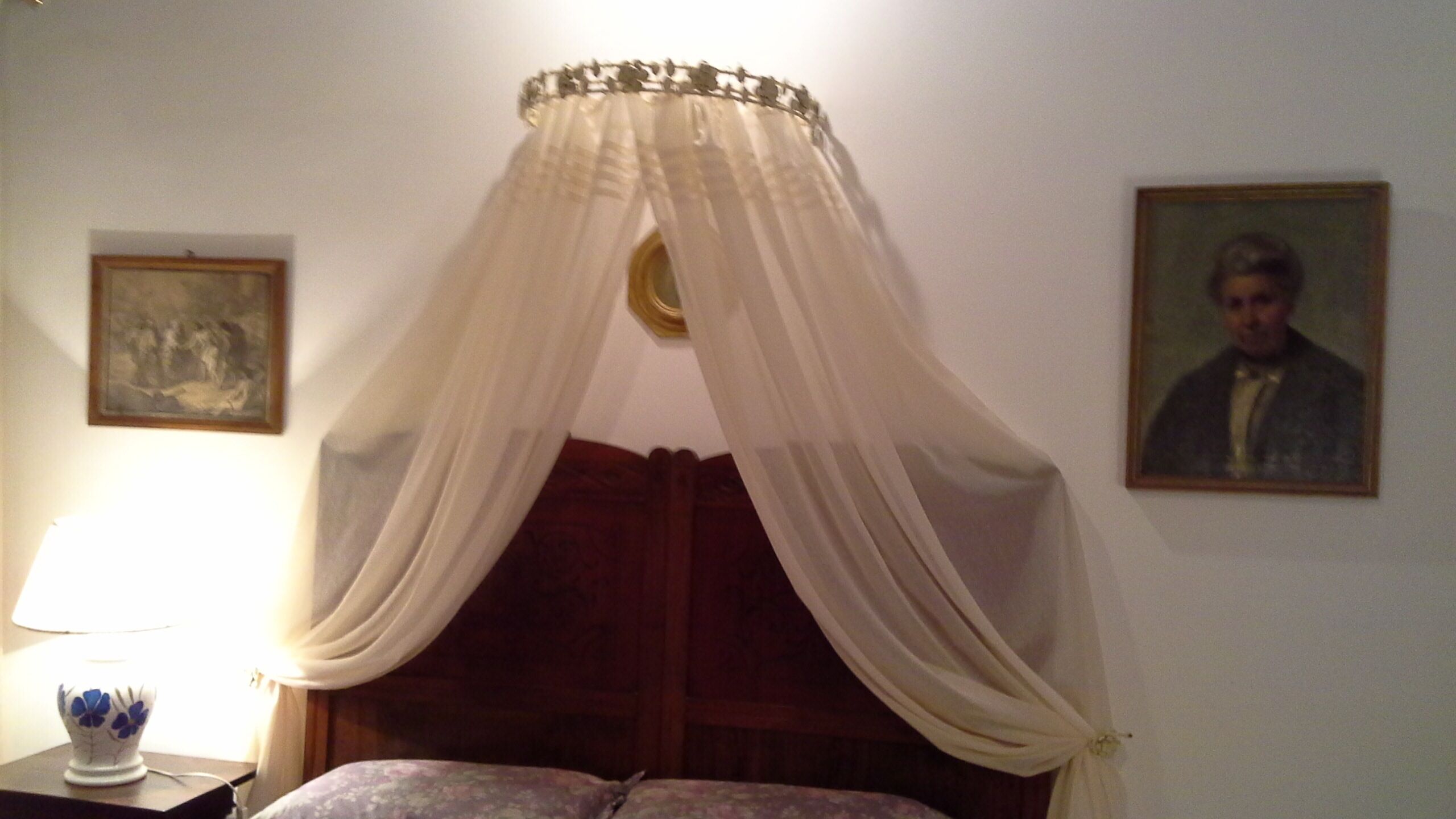 A bedroom with a canopy bed, a bedside table with a lamp, and two framed pictures on the wall.