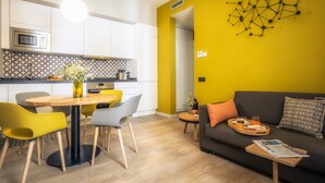 Une cuisine moderne avec des armoires blanches, un four à micro-ondes et un crédence décorée. On y trouve un coin repas avec une table et des chaises en bois, ainsi qu’un salon avec un canapé et une table basse.