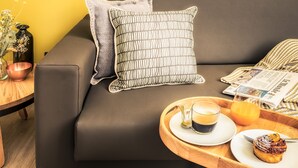 Un salon moderne avec un canapé foncé, une table d’appoint en bois, un plateau sur lequel se trouvent une tasse de café, un verre de jus d’orange et une assiette avec une pâtisserie.