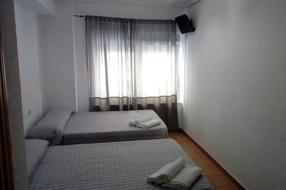 A small, clean room with two beds, a flat-screen TV mounted on the wall, and a window with sheer curtains.