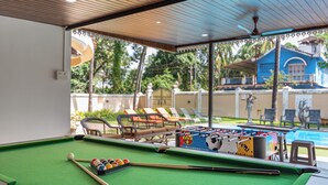 A pool table with a foosball table, pool cues, and balls, set in a covered area with a view of a pool and lounge chairs.