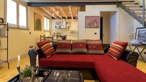 Ein modernes Wohnzimmer mit einem roten Ecksofa, einem schwarzen Lederhocker und einem Glastisch. Im Hintergrund ist ein Essbereich mit einer sichtbaren Küche zu sehen.