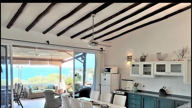 Une cuisine moderne avec des meubles blancs, un coin repas avec des chaises blanches et une vue sur l’océan grâce à de grandes portes vitrées.